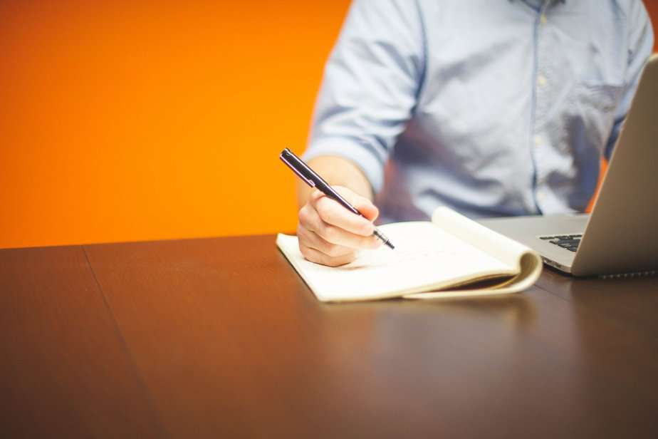 Ein Mann sitzt vor einer leuchten orangeroten Wand und macht sich am Computer Notizen.